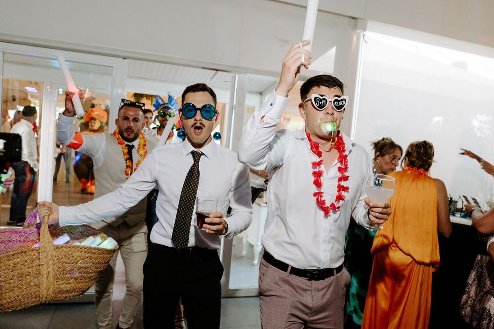 Men in novelty sunglasses and party accessories celebrate at a festive indoor wedding reception party.