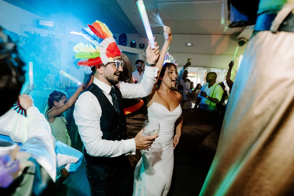 Bride and groom dancing joyfully at wedding reception, groom wearing colorful feathered hat, guests celebrating in background.