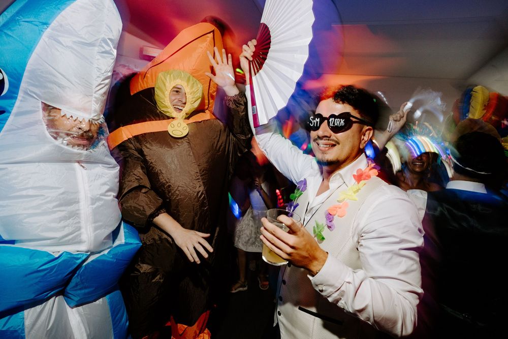 Person in white suit with sunglasses poses next to inflatable alien costume at a colorful costume party.