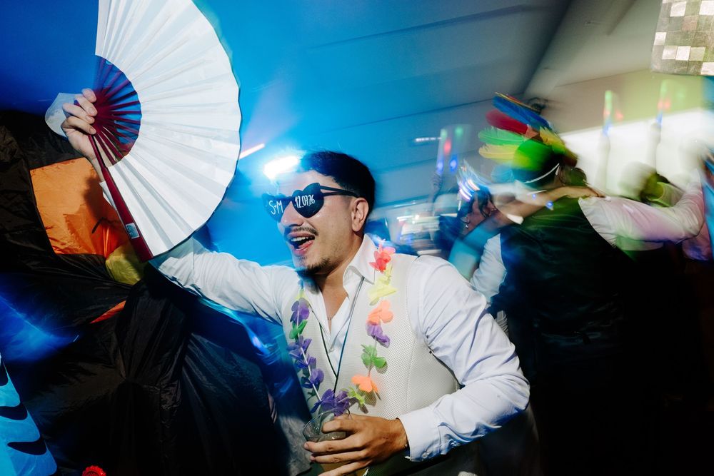 Man in sunglasses holding white fan wearing floral lei dancing at a lively party celebration.