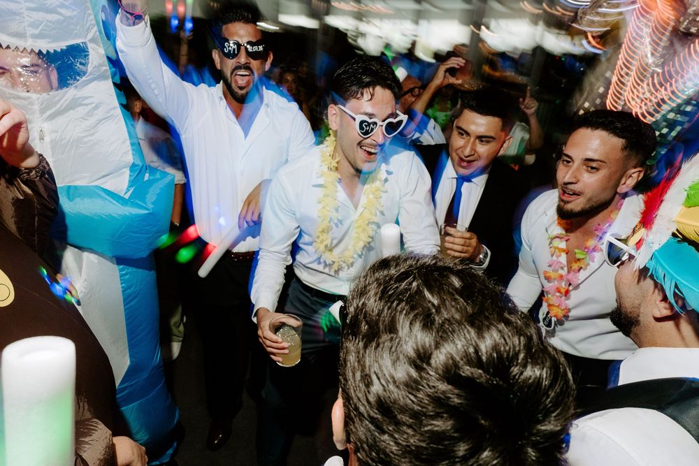 Group of young men dancing and celebrating at a lively party under a tent with colorful decorations.