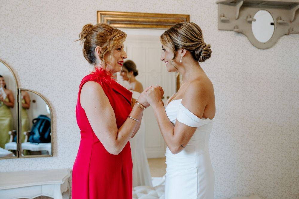 Bride in white off-shoulder gown shares a joyful moment with woman in red dress during wedding preparations.