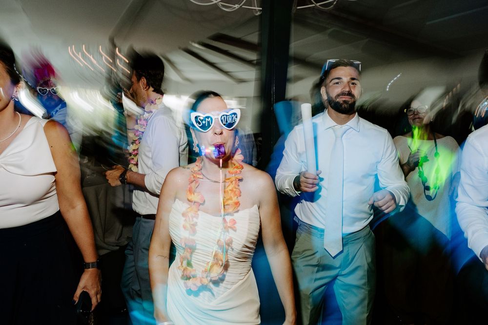 Bride wearing heart sunglasses and floral lei dances at lively wedding reception party with guests.