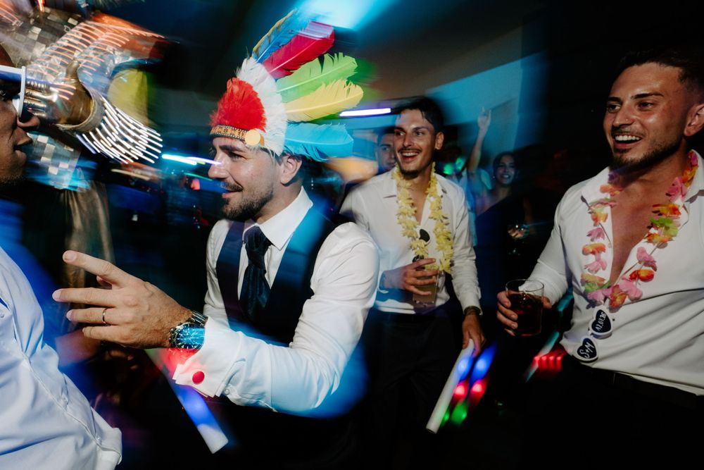 Men in formal wear with colorful party hats and LED accessories dancing at a wedding reception.