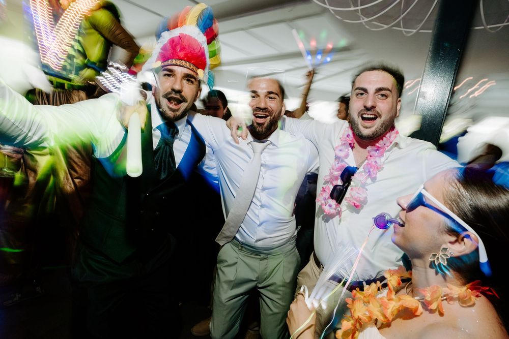 Three men in suits celebrating with glow sticks and colorful accessories at a festive wedding party.