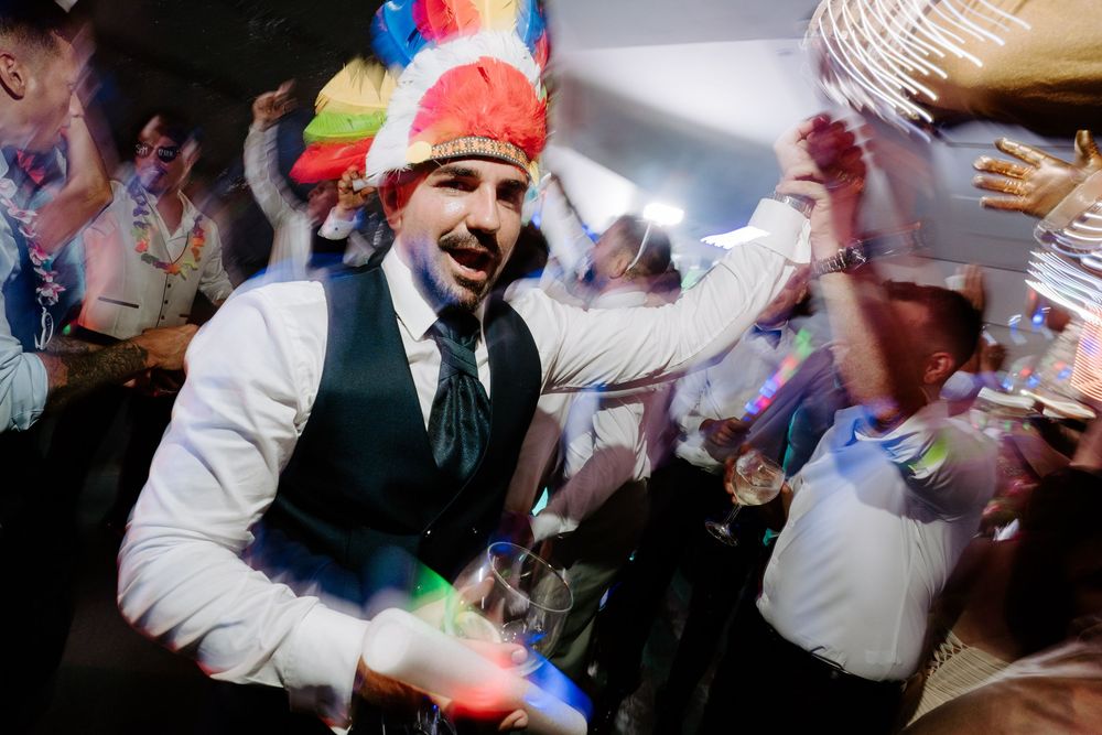 Man in white shirt and vest wearing colorful feathered hat dancing at a lively party celebration.