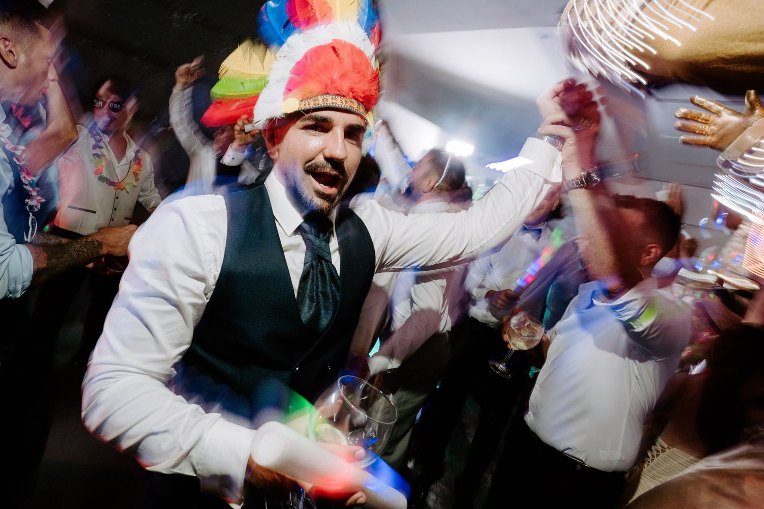 Man in white shirt and vest wearing colorful feathered hat dancing at a lively party celebration.