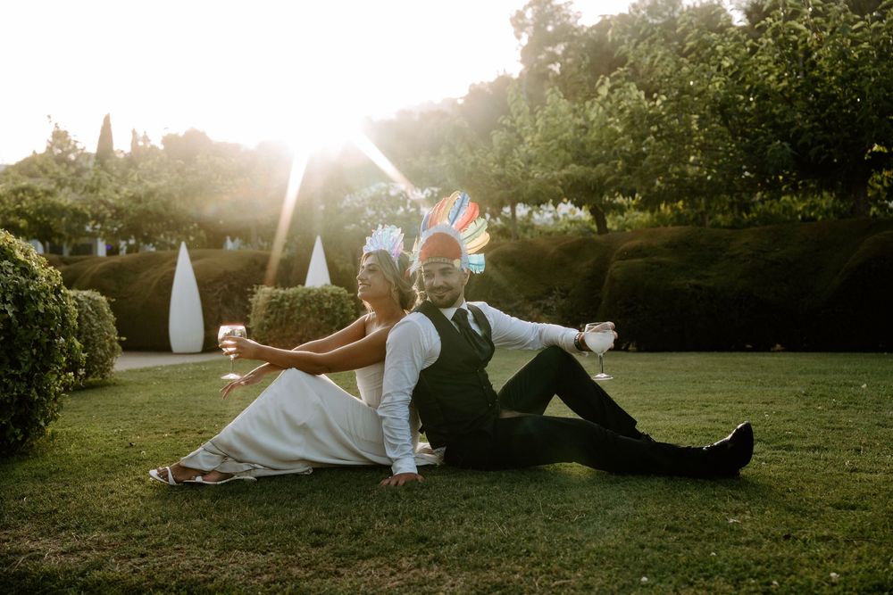 Bride and groom sitting back-to-back on grass holding drinks and fun props in golden sunset garden.