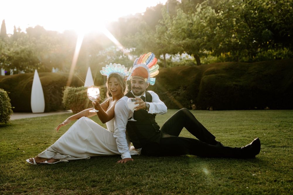 Happy couple sitting on lawn with colorful props and drinks bathed in warm golden sunlight outdoors.