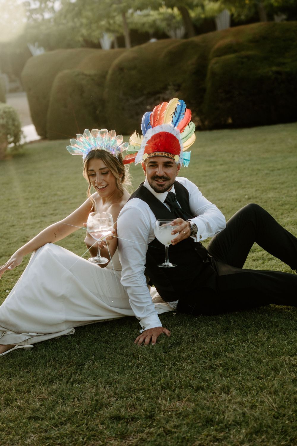 Bride and groom sitting on grass wearing colorful feathered headdresses while holding wine glasses at outdoor wedding.