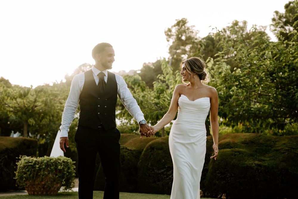 Bride and groom holding hands walking through lush garden in golden sunset light, bride in strapless white gown.