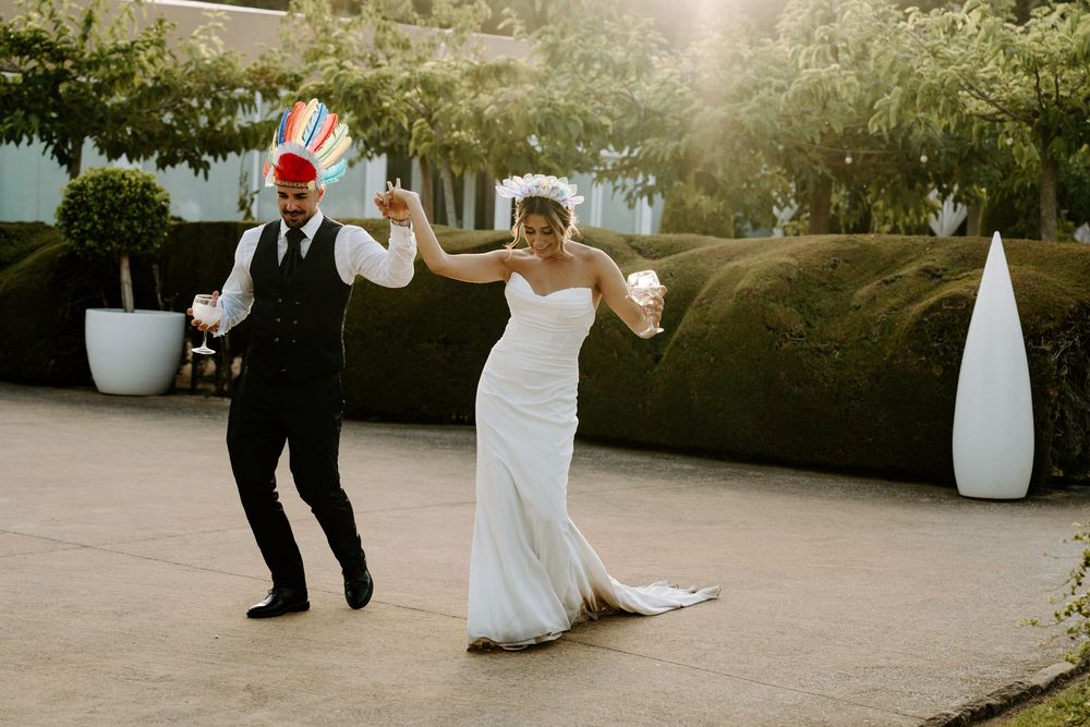 A joyful bride and groom dance outdoors in a garden setting, with the groom wearing a colorful hat on their wedding day.