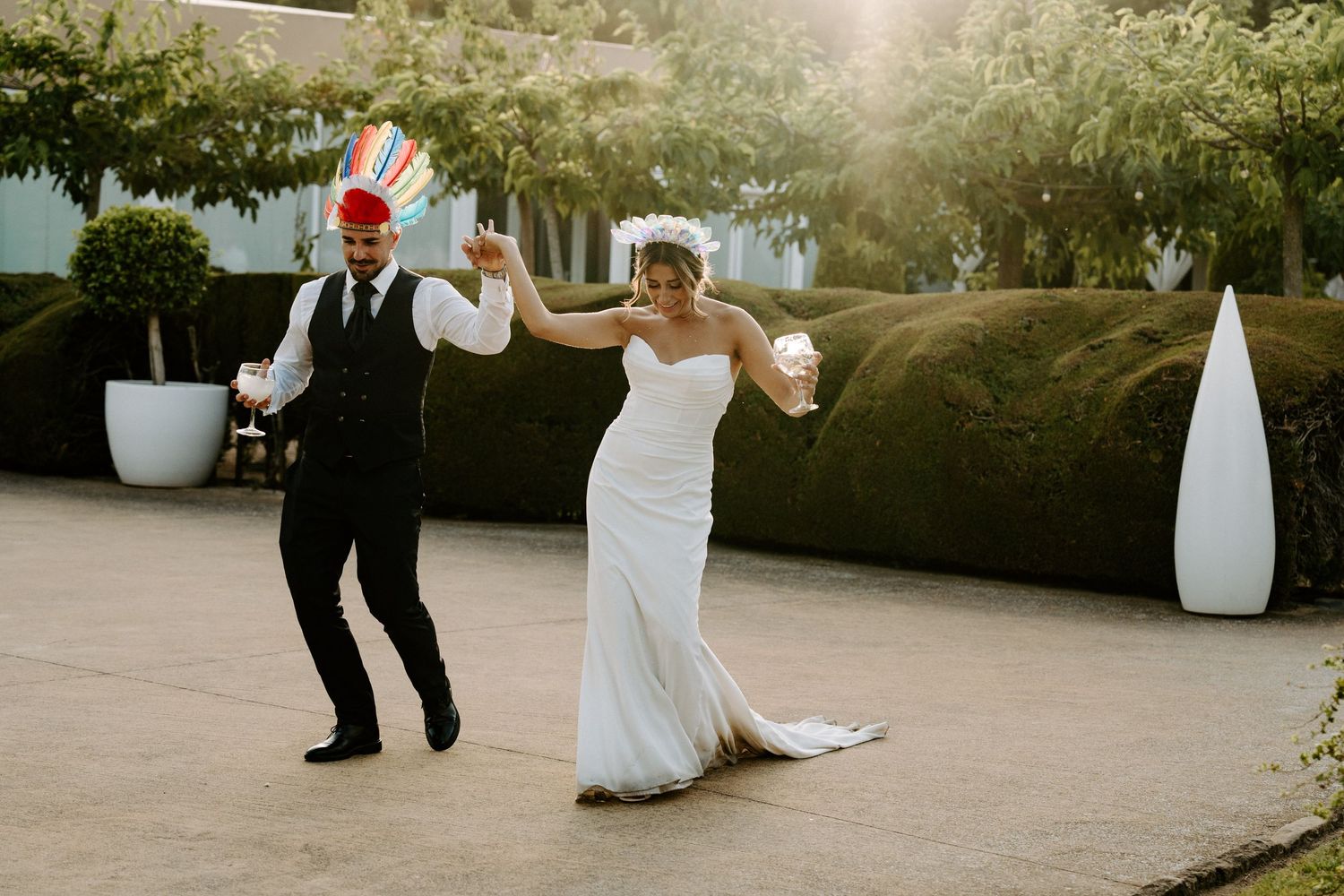 A joyful bride and groom dance outdoors in a garden setting, with the groom wearing a colorful hat on their wedding day.
