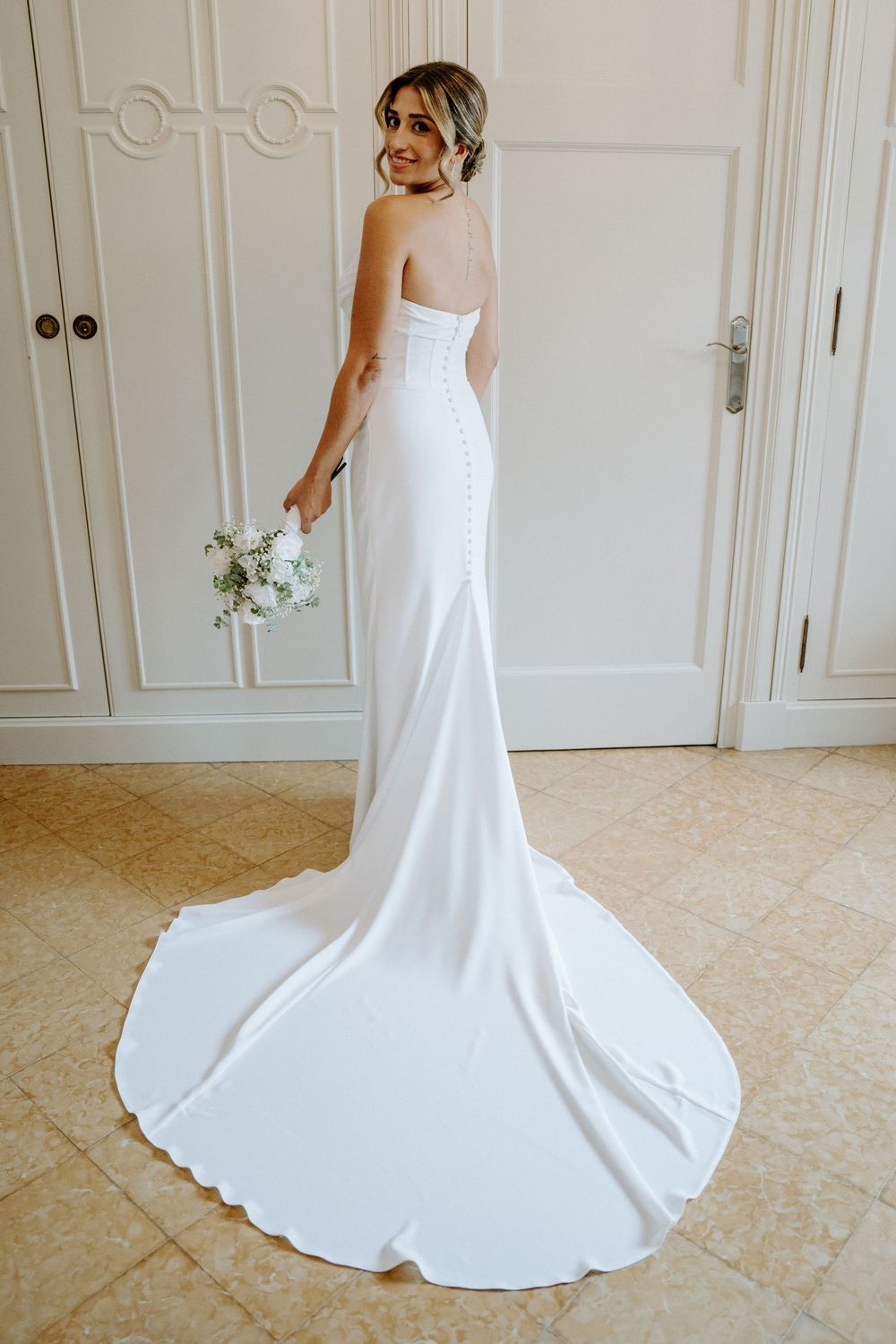 Elegant bride in strapless white fitted gown with button back train holds bouquet indoors.