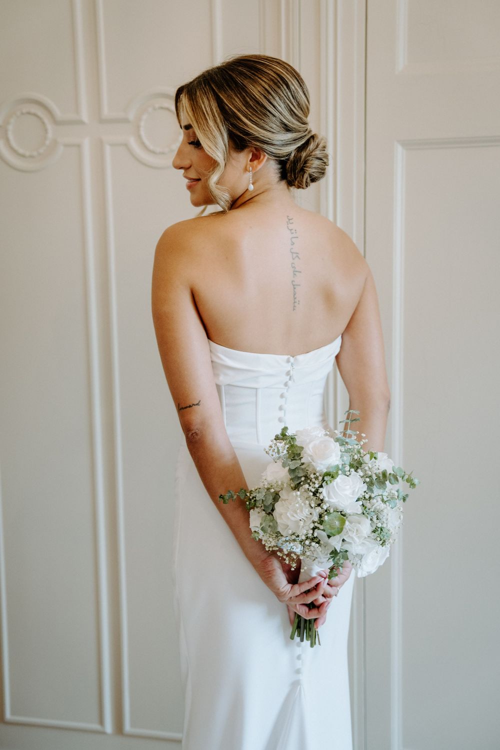 Bride in strapless white gown holding bouquet of white roses, showing back tattoo and button detail.