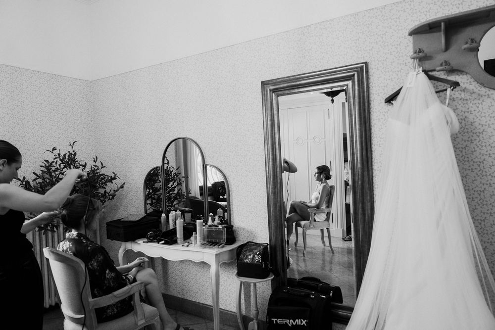 Black and white photo of bride getting hair done before wedding, with gown hanging and mirror reflection in elegant room.