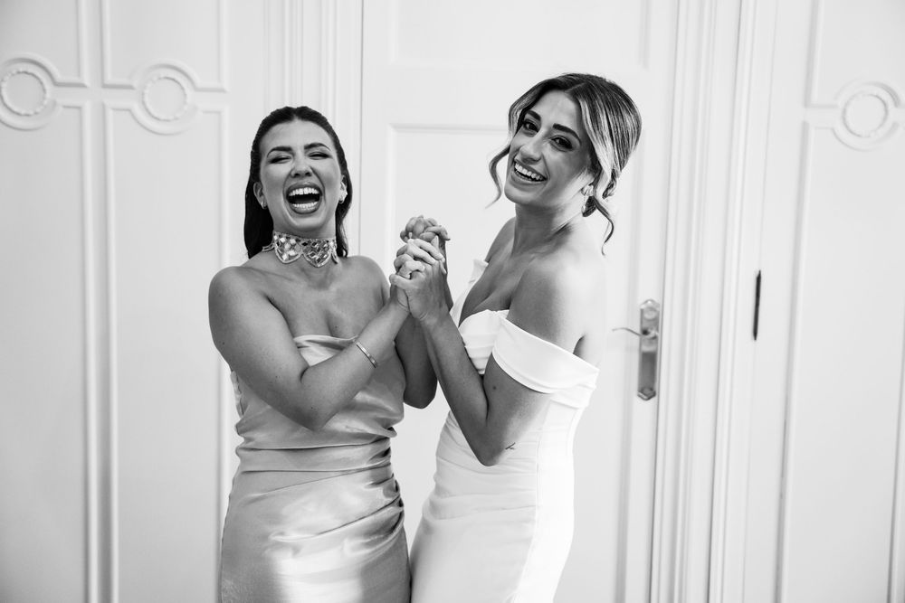Two elegantly dressed women laughing and holding hands in black and white photo, one in bridal gown.