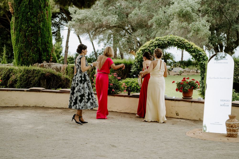 Four elegantly dressed women socializing outdoors at a wedding venue surrounded by lush greenery and cypress trees.