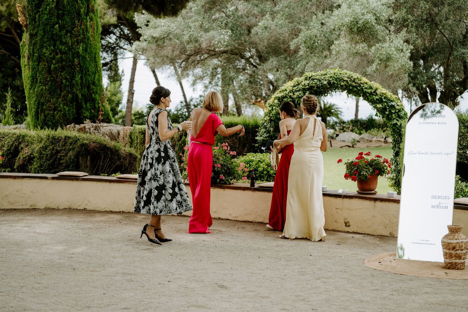 Four elegantly dressed women socializing outdoors at a wedding venue surrounded by lush greenery and cypress trees.