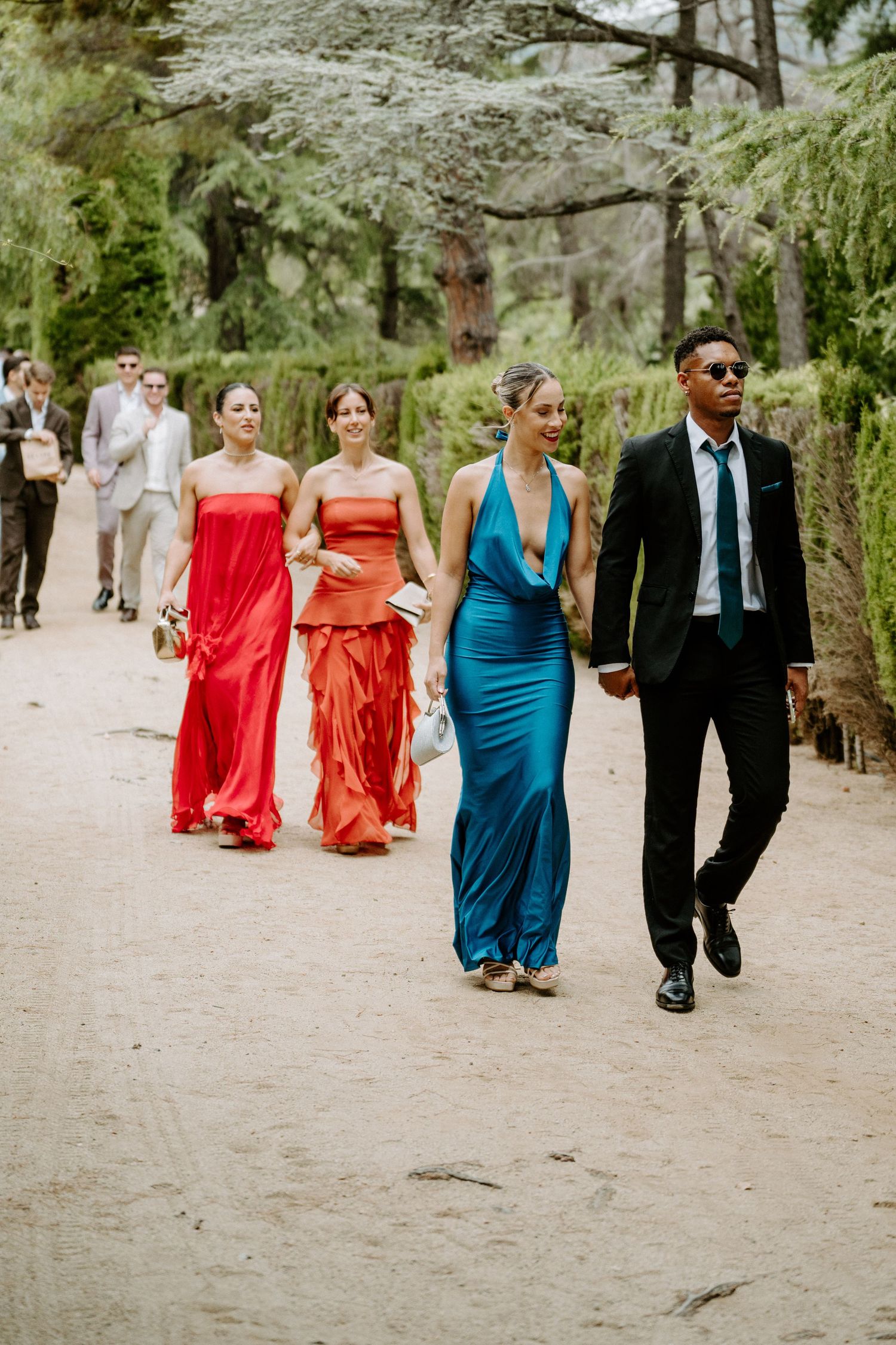 Wedding guests walk through tree-lined garden path, bridesmaids in red and teal satin gowns.