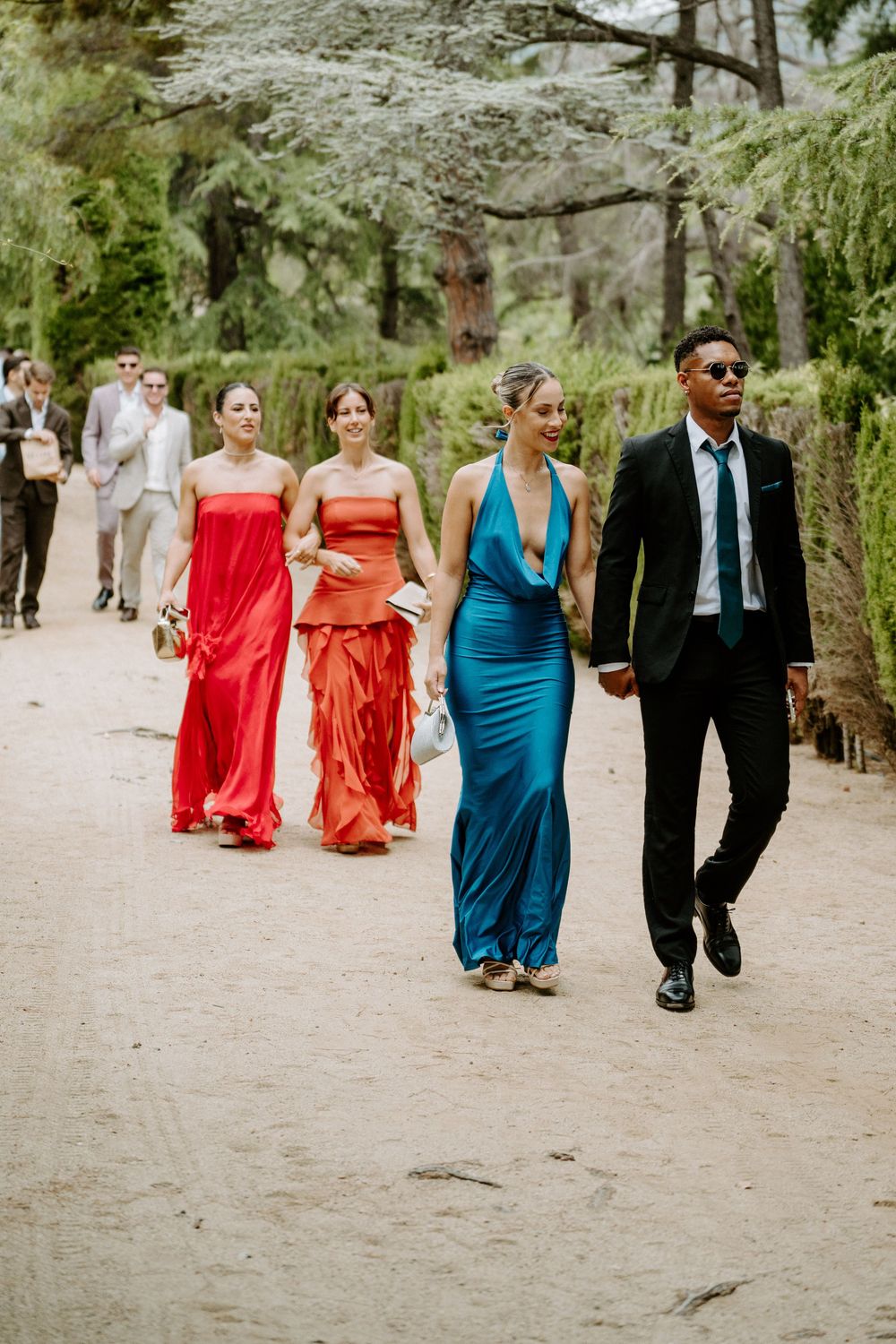 Wedding guests walk through tree-lined garden path, bridesmaids in red and teal satin gowns.