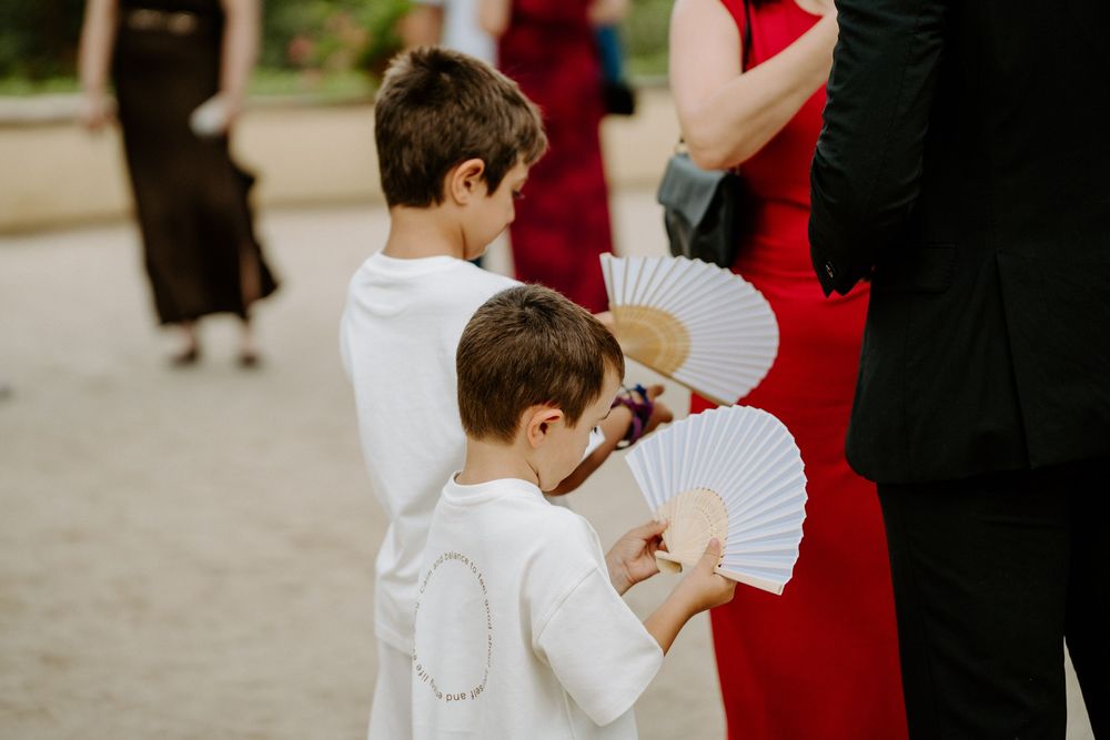 Two young boys in white outfits holding decorative hand fans at an outdoor wedding celebration.