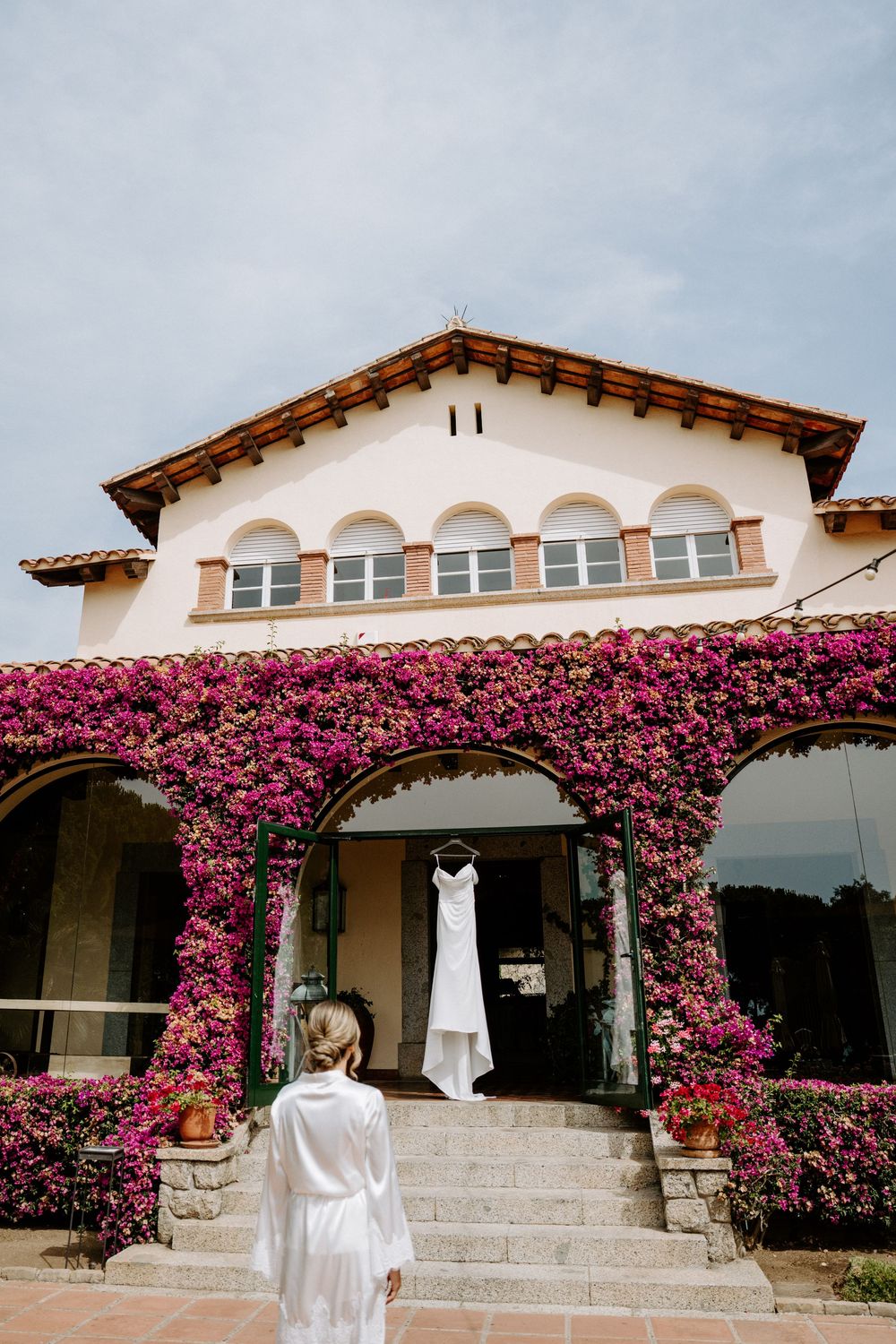 Bride in white robe stands before a Spanish-style villa covered in vibrant pink bougainvillea with wedding dress hanging in archway.