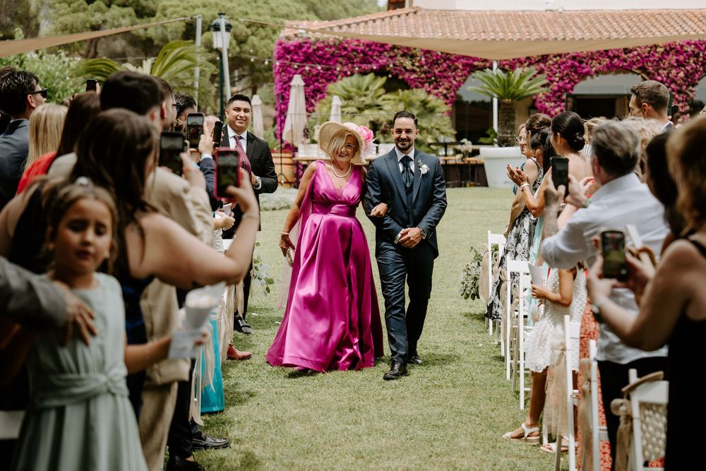 Groom escorts woman in pink gown down outdoor wedding aisle lined with guests and vibrant fuchsia flowers.