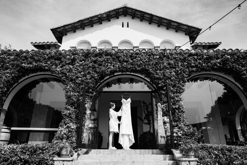 Black and white photo of a bride holding her wedding dress in an ivy-covered arched building entrance.