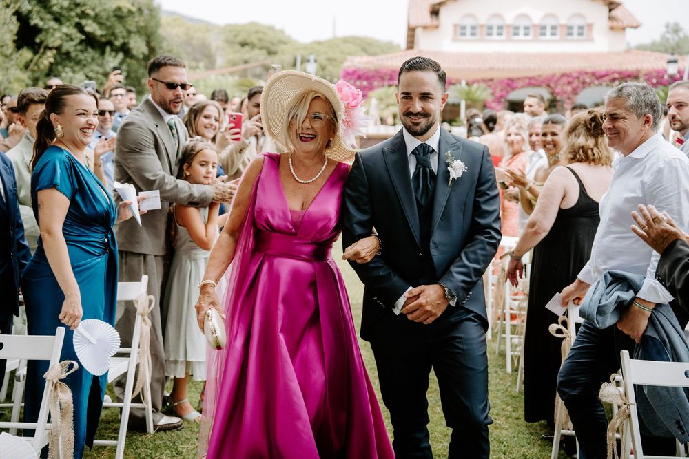 Groom escorts elegantly dressed woman in pink gown down wedding aisle surrounded by cheering guests outdoors.