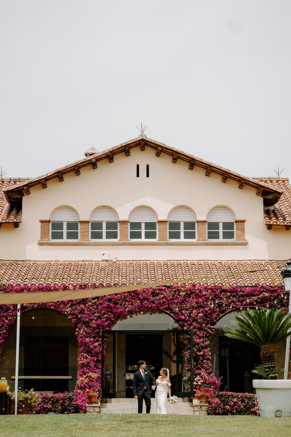 Bride and groom stand before stunning Spanish-style villa adorned with vibrant bougainvillea flowers.