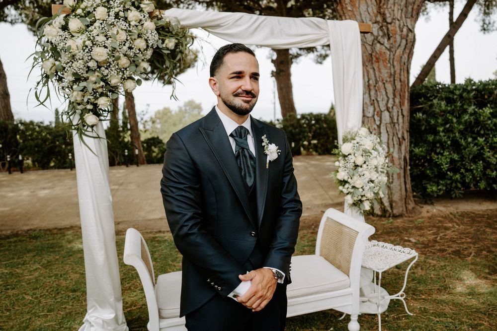 Groom in navy suit smiling at outdoor wedding ceremony beneath floral arch with white draping.