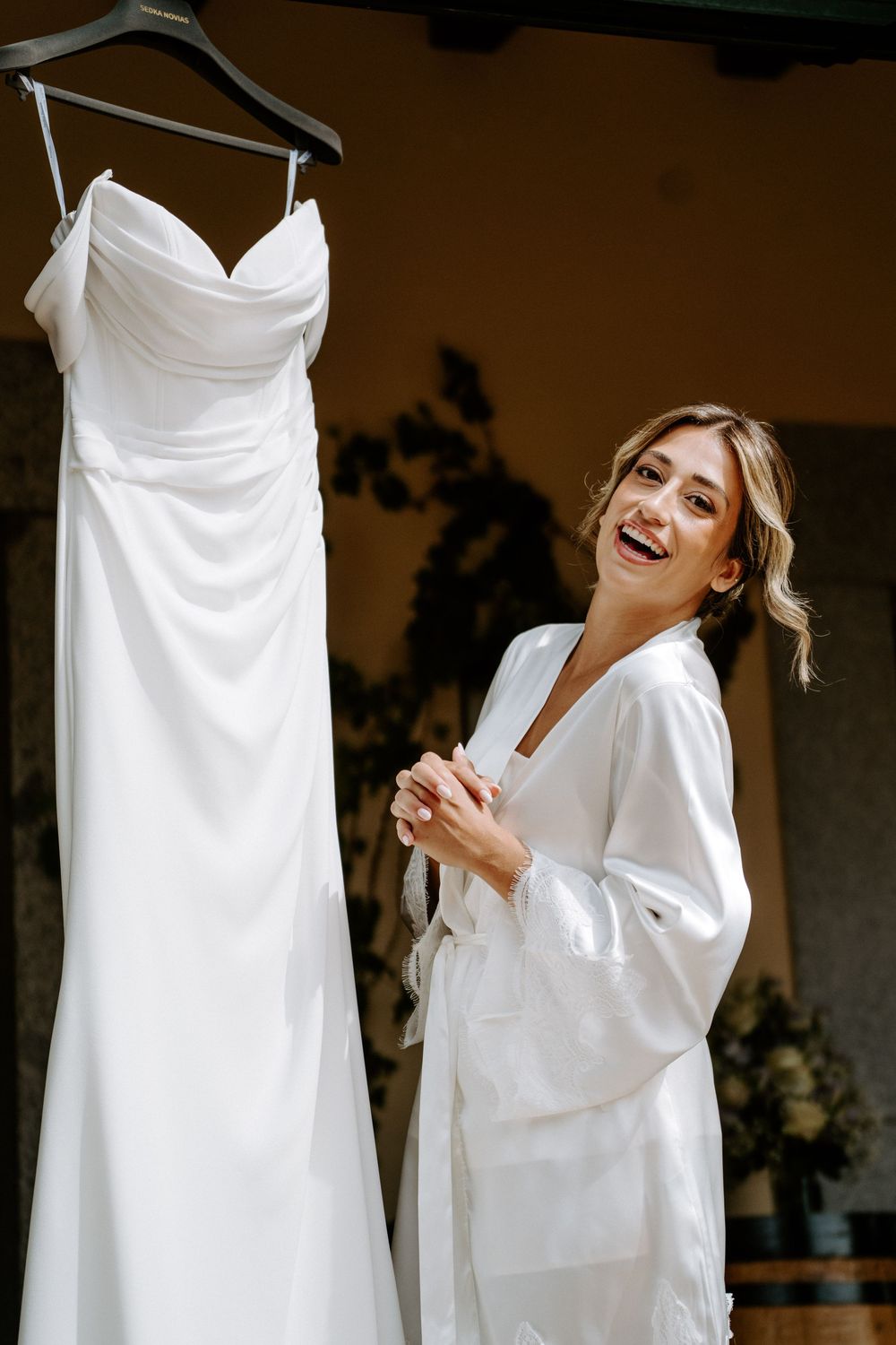 Smiling bride in white robe standing next to her elegant wedding dress on a hanger before the ceremony.