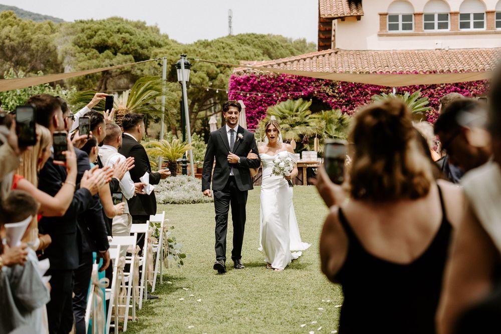 Newlyweds walk down outdoor aisle as guests applaud at a sunny garden wedding ceremony.