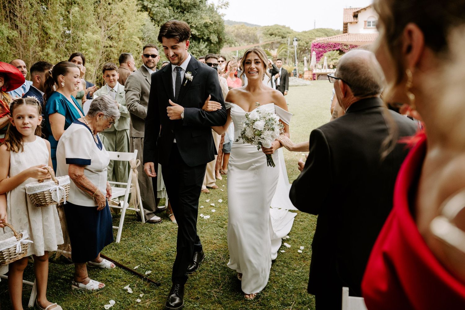 Bride and groom walking down outdoor aisle surrounded by cheering wedding guests on sunny day.