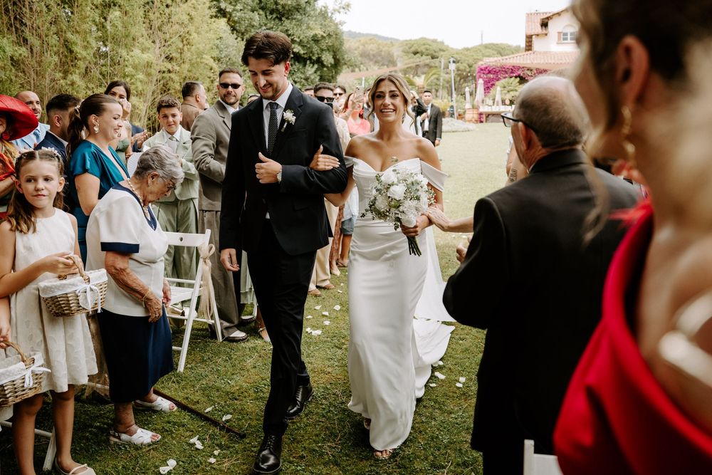 Bride and groom walking down outdoor aisle surrounded by cheering wedding guests on sunny day.