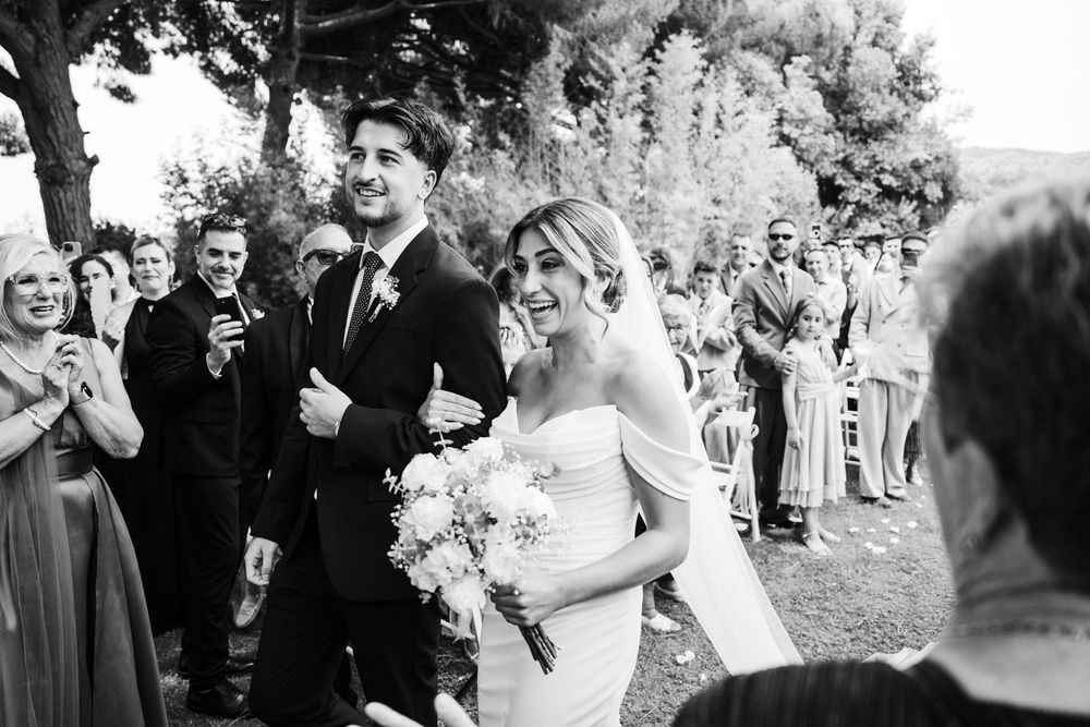Black and white photo of smiling bride and groom walking back down aisle after outdoor wedding ceremony.