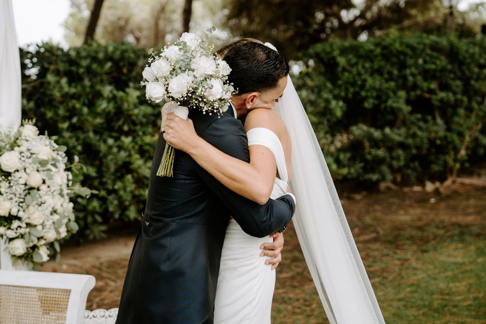 Bride and groom share an emotional embrace outdoors during wedding ceremony, bride holding white bouquet wearing veil.