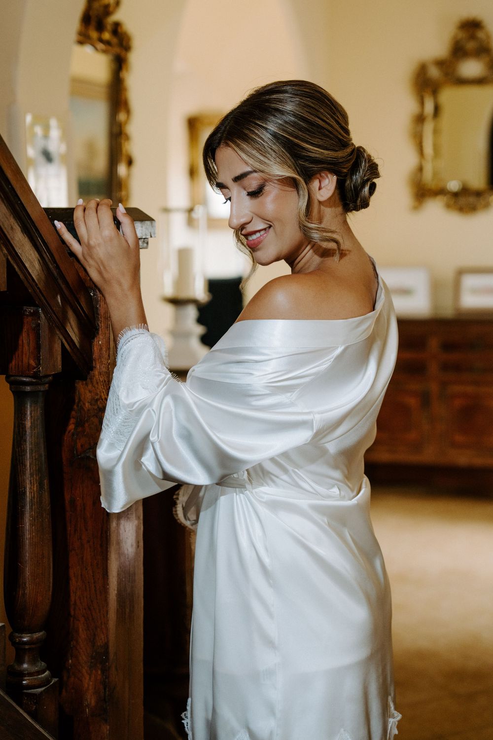Smiling bride in white satin robe standing by wooden staircase in elegant room with ornate gold mirrors.
