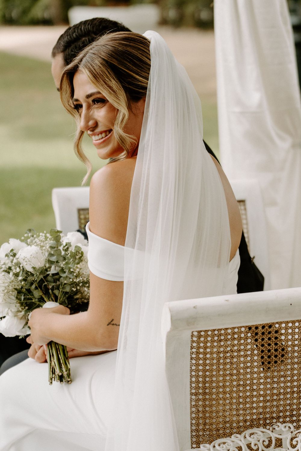 Smiling bride in white off-shoulder gown and veil holds bouquet while seated outdoors at wedding ceremony.