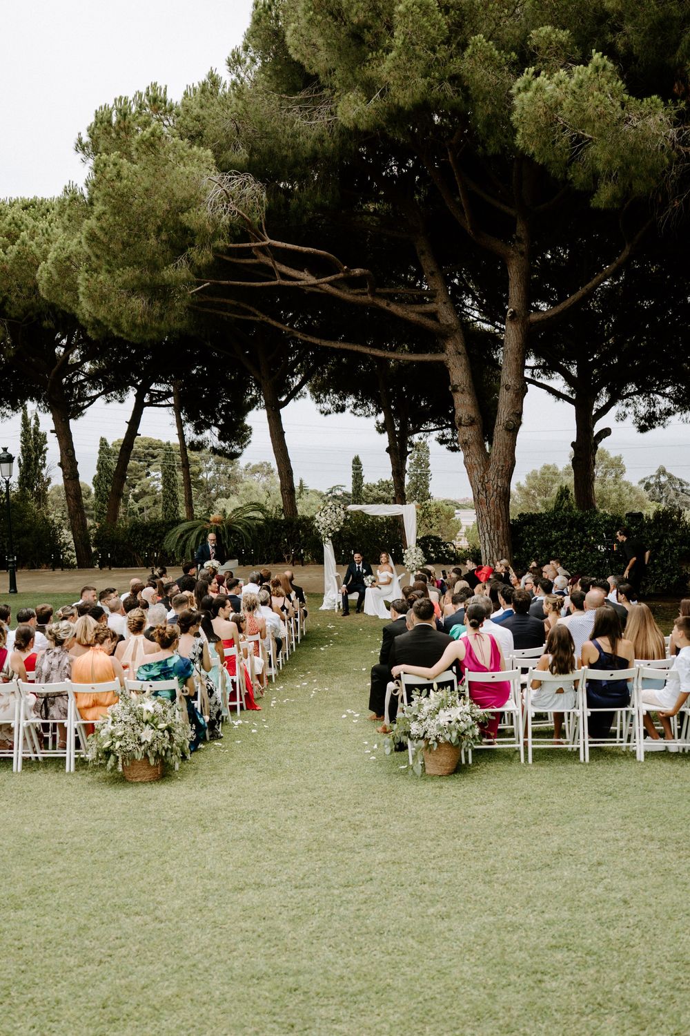 Outdoor wedding ceremony on green lawn with guests seated in white chairs beneath tall pine trees.
