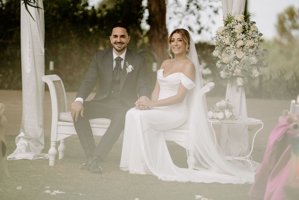 A smiling bride in a white off-shoulder gown and groom in a dark suit sit together during an outdoor wedding ceremony.