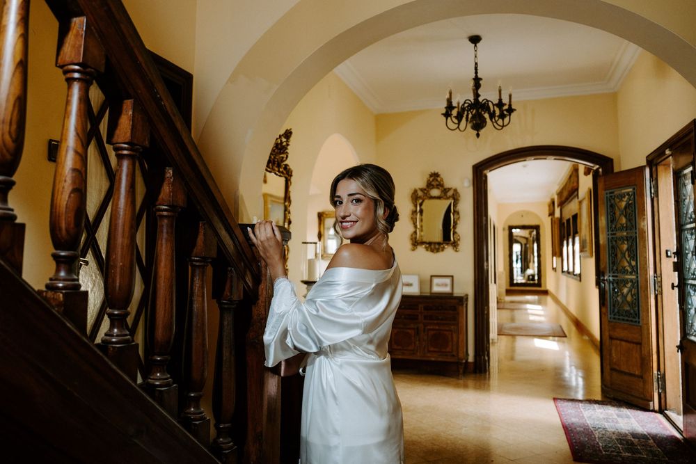 Elegant bride in white satin gown standing on wooden staircase in luxurious villa hallway with chandelier.