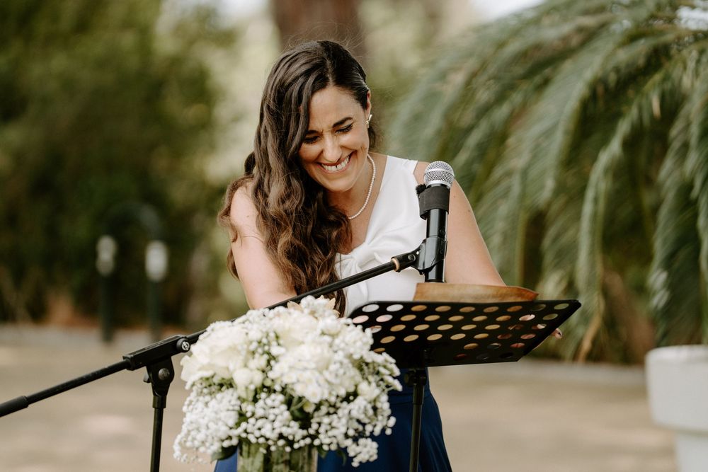 Smiling woman in white top reading at a microphone podium decorated with white flowers at a wedding.