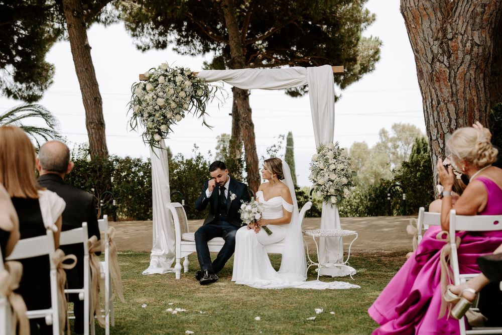 Bride and groom seated at outdoor wedding altar under white draped arch surrounded by floral arrangements.