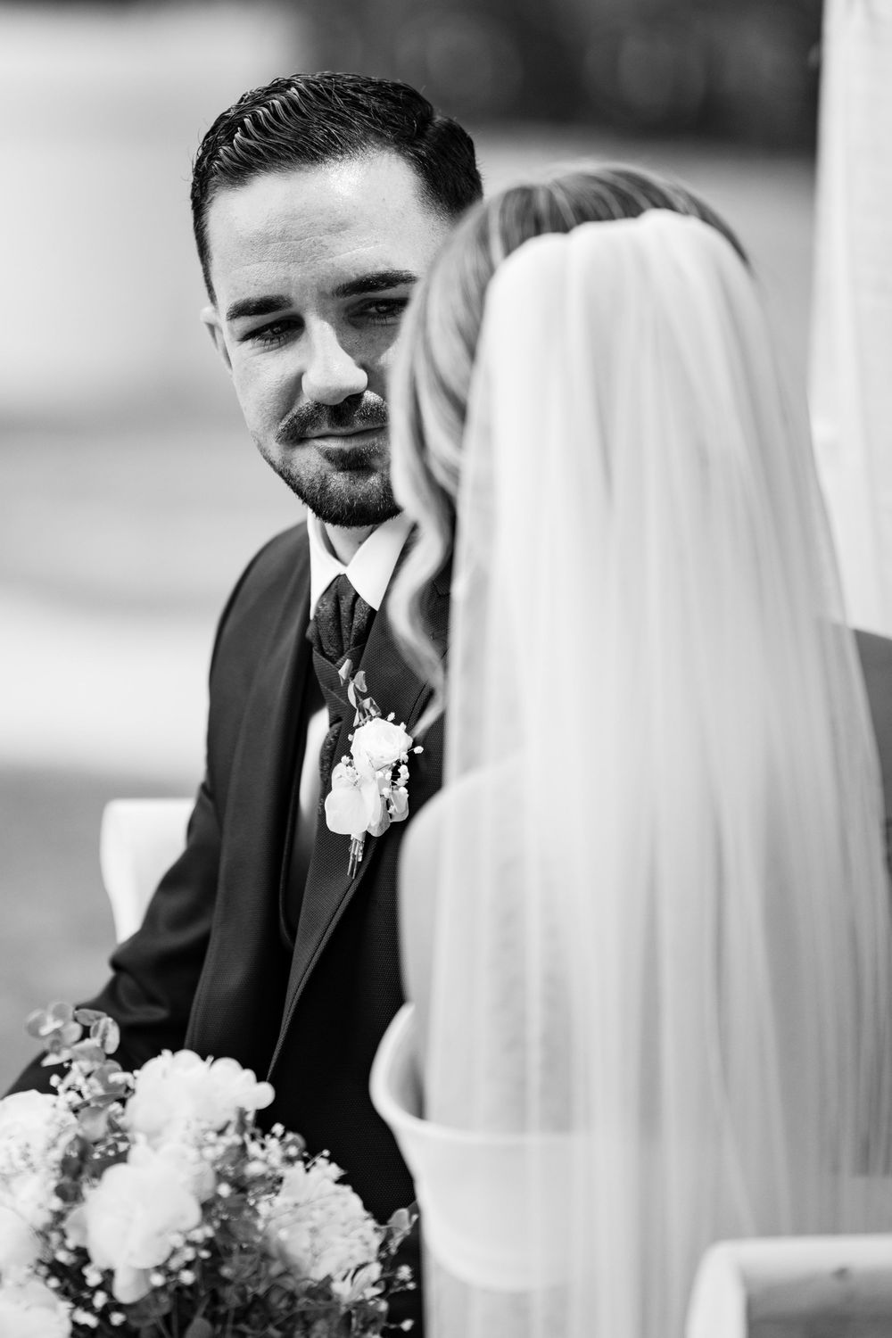 Black and white photo of groom gazing lovingly at bride during outdoor wedding ceremony.