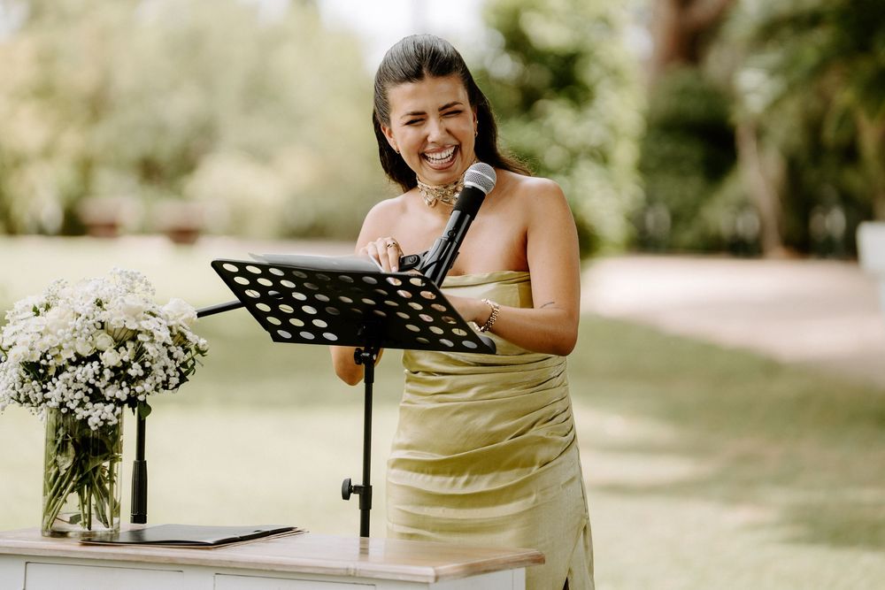 Smiling female officiant in gold dress speaks into microphone at outdoor wedding ceremony podium.