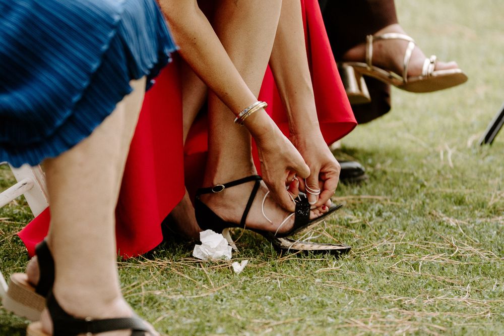 Wedding guests adjust strappy heeled sandals on green grass during outdoor ceremony.