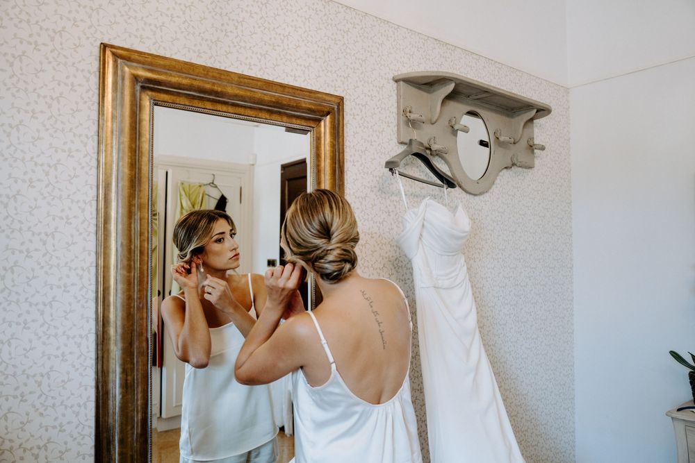 Bride in white slip dress adjusts earrings before large gold mirror with wedding gown hanging on wall hook.