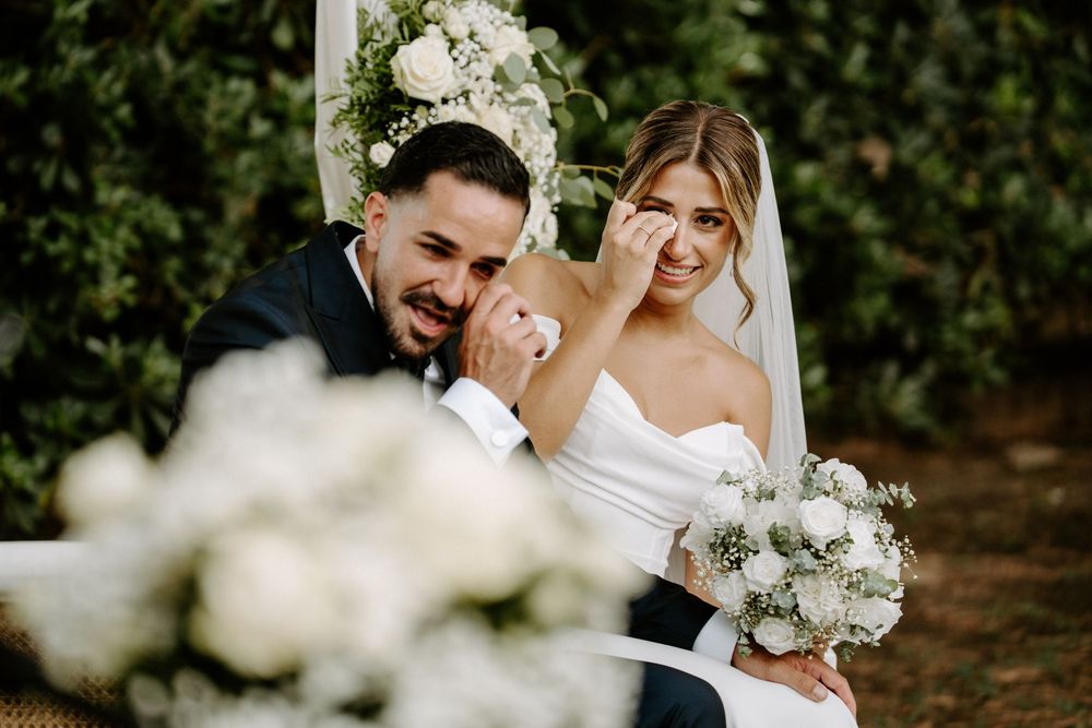 Emotional bride wiping tears while groom smiles during outdoor wedding ceremony with white floral bouquet.